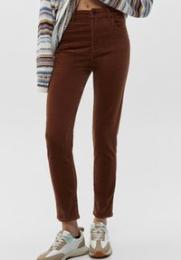 Jean en velours côtelé marron avec une coupe slim, présentant une taille haute et des poches à l'avant, associés à des baskets blanches et beiges.