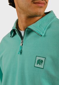 Maglietta con zip di colore turchese, caratterizzata da un colletto alto, una texture morbida e un logo di elefante ricamato di colore verde sul lato sinistro del petto.