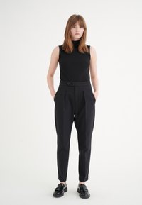 Haut sans manches noir avec une texture côtelée associé à un pantalon noir taille haute avec des plis et un détail de bouton. Des mocassins noirs complètent la tenue.