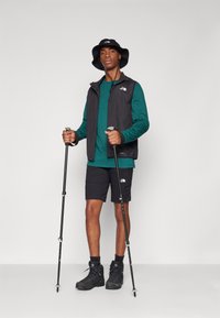 Sort vest over en teal langærmet skjorte, sorte shorts og sorte vandrestøvler. Holder på vandrestave, iført en sort bøttehat.
