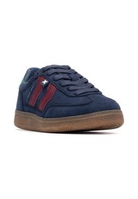 Zapatillas de ante azul marino con rayas burdeos, acento verde en el talón y suela de goma marrón texturizada. Presenta un diseño de cordones y un detalle de logo.