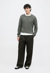 Sivi teksturirani pulover s okruglim izrezom, preko bijele košulje. Labave tamnosmeđe hlače, crno-bijele cipele. Jednostavna pozadina.