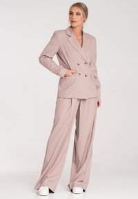 Jasnoróżowy garnitur w prążki z podwójnym zapięciem na marynarce z złotymi guzikami oraz szerokimi spodniami. Gładki materiał, dopasowany krój i kieszenie.
