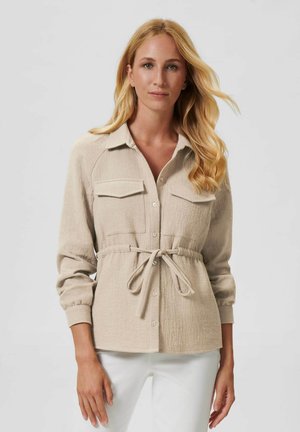 Beige knoopshirt met twee borstzakken, een getailleerde taille en lange mouwen. Textuurstof en strikdetail verbeteren het ontwerp.