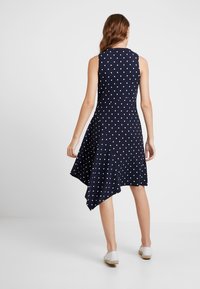 Námorně modré bezramenní šaty s bílými puntíky, rozšířený lem, asymetrický design, vyrobené z lehké látky. Žádné viditelné hardwarové prvky.