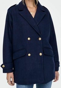 Cappotto doppiopetto blu navy con bottoni dorati, tasche a patta e spalline, indossato sopra jeans blu.