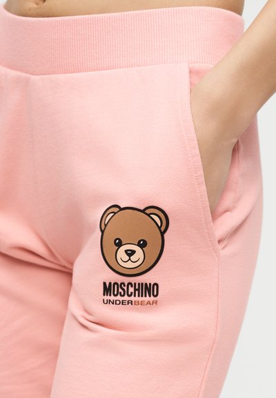 Κοντινή λήψη τσέπης ροζ φόρμας με ένα χέρι μέσα, που διαθέτει λογότυπο με πρόσωπο καφέ αρκούδας και κείμενο "MOSCHINO UNDERBEAR" από κάτω.