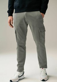 Graue Cargo-Hosen mit Seitentaschen, tailliertem Design und glatter Stoffstruktur. Kombiniert mit weißen Sneakers mit schwarzen und grauen Akzenten.
