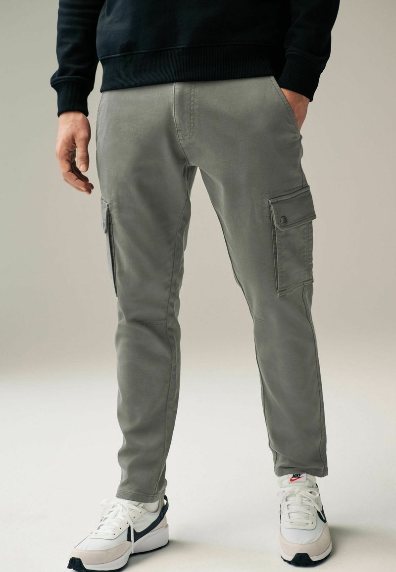 Graue Cargo-Hosen mit Seitentaschen, tailliertem Design und glatter Stoffstruktur. Kombiniert mit weißen Sneakers mit schwarzen und grauen Akzenten.