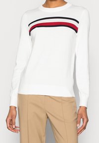 Maglione bianco con scollo rotondo, caratterizzato da una striscia orizzontale testurizzata in rosso, bianco e nero sul petto. Tessuto in maglia morbida.
