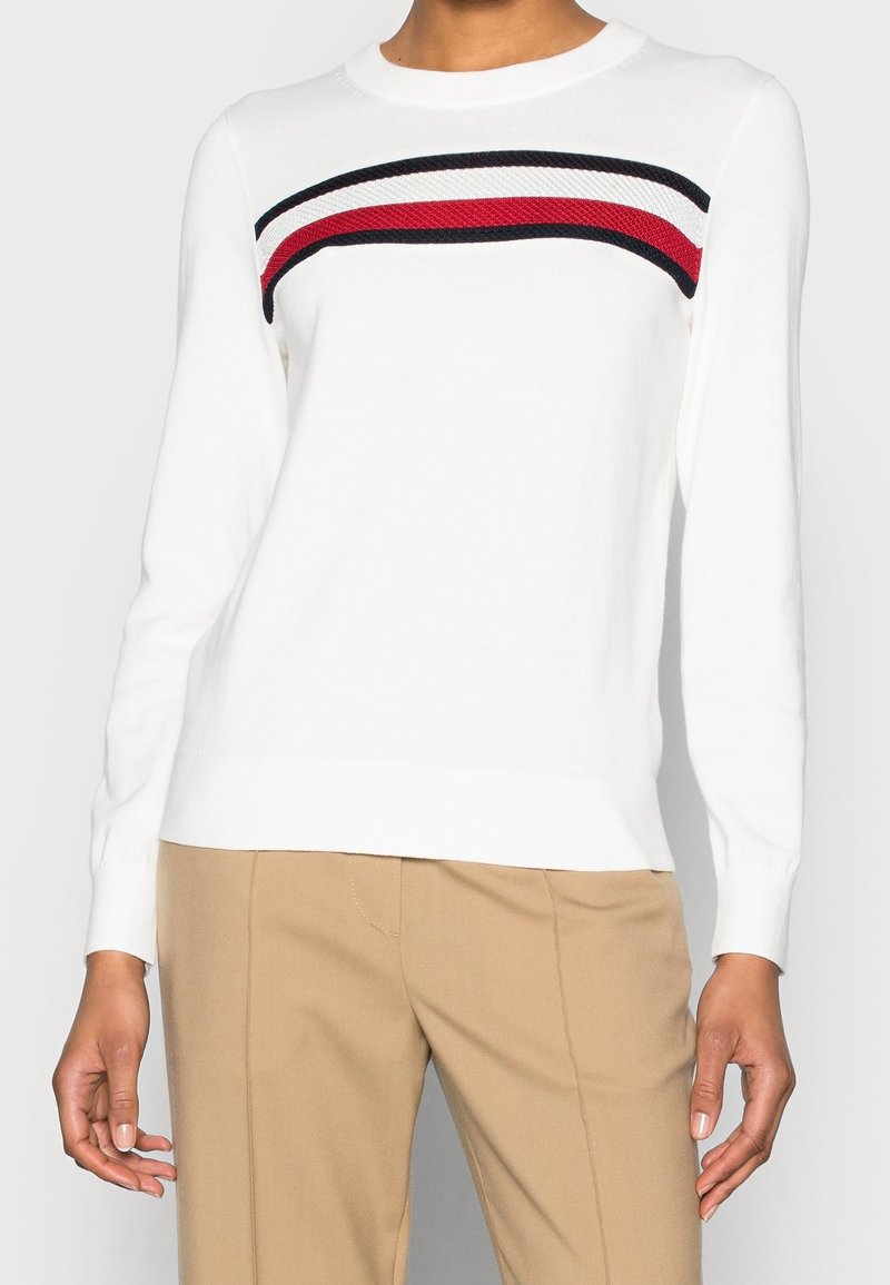 Maglione bianco con scollo rotondo, caratterizzato da una striscia orizzontale testurizzata in rosso, bianco e nero sul petto. Tessuto in maglia morbida.