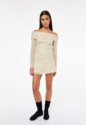 Giovane donna con un vestito beige scollato con piccoli pois neri, calzini neri e scarpe nere, in piedi su sfondo bianco.