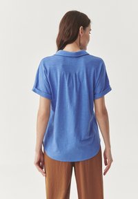 Camicia blu a maniche corte con colletto, caratterizzata da maniche arrotolate, una piega sulla schiena e un orlo stondato. Realizzata in tessuto leggero e testurizzato.