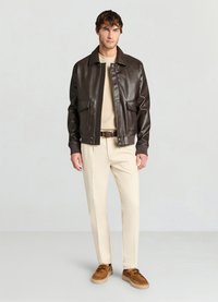 Veste aviateur en cuir marron avec fermeture éclair à l'avant, deux grandes poches avant, portée sur un pull beige à col rond, associée à un pantalon crème et des chaussures marron.