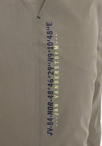Light gray shirt featuring bold dark blue and yellow text. Notable details include geographic coordinates and designer name on the side.