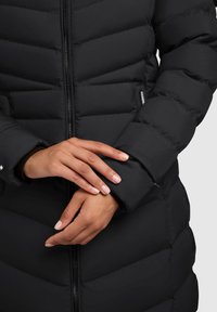 Zwarte pufferjack met een quiltpatroon, voorzien van een ritssluiting en elastische manchetten. De stof lijkt glad en weersbestendig.