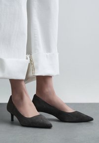 Sivé semišové lodičky so špicatou špičkou a nízkym opätkom, skombinované s bielymi nohavicami so strapcami na leme, predstavujú jednoduchý a elegantný dizajn.