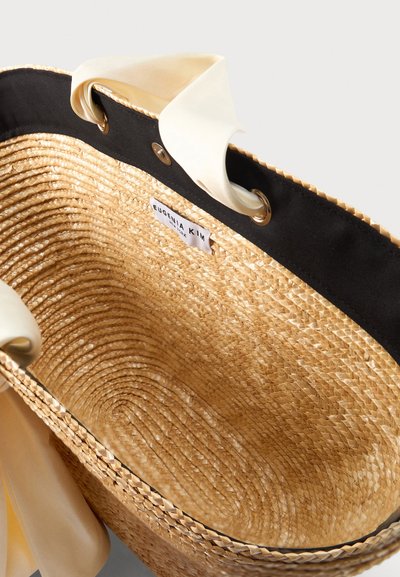 Sac cabas en paille tissée avec bordure en tissu noir, poignées en satin beige passées dans des œillets métalliques, et étiquette « Eugenia Kim » à l'intérieur.