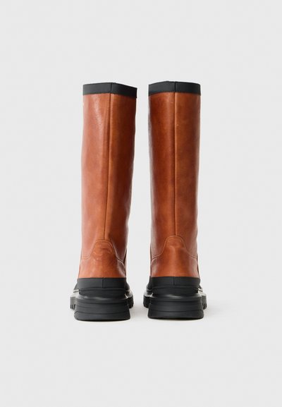 Καφέ δερμάτινα μπότες μέχρι το γόνατο με λεία επιφάνεια, μαύρες καουτσούκ σόλες και αντιθετικό μαύρο επάνω χείλος, με απλές λεπτομέρειες ραφής.