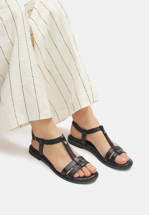 Czarne skórzane sandały z projektem w kształcie litery T, krzyżującymi się paskami i płaską podeszwą. Paski mają gładką teksturę i zapięcie na klamrę.