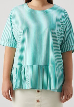 Femme portant un chemisier ample à manches courtes rayé verticalement turquoise et blanc, avec un ourlet peplum froncé, et une jupe blanche boutonnée.
