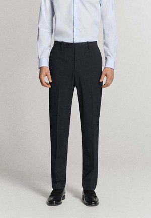 Mężczyzna ubrany w czarne eleganckie spodnie, jasnoniebieską koszulę na guziki i czarne eleganckie buty, stojący na jednolitym jasnym tle.