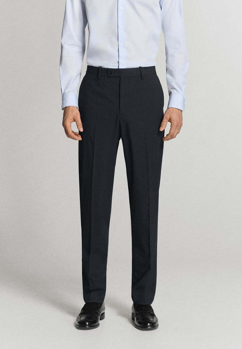 Homme portant un pantalon habillé noir, une chemise bleu clair boutonnée et des chaussures habillées noires, debout devant un fond clair uni.
