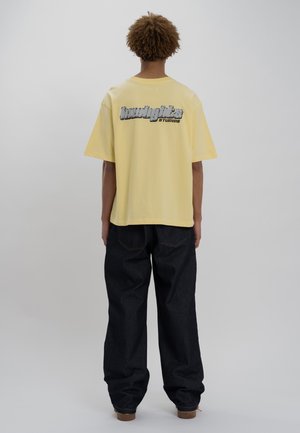 T-shirt en coton jaune avec un logo graphique au dos, associé à un jean en denim bleu foncé à coupe ample. La texture semble douce et décontractée.
