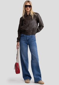 Giacca bomber in pelle marrone con vita elastica, abbinata a jeans in denim blu a gamba larga. Tieni una piccola borsa a tracolla rossa.