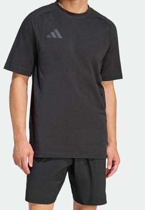 Homme portant un t-shirt de sport noir à manches courtes avec un logo discret sur la poitrine et un short noir assorti, debout les bras détendus.