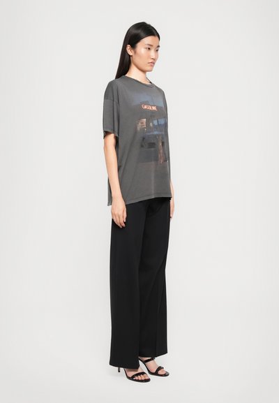 O tânără poartă pantaloni negri cu croială largă, sandale negre cu toc și un tricou gri închis, lejer, cu imprimeu de benzinărie.