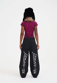 Pantalones de mezclilla anchos negros con letras blancas en los laterales, combinados con una camiseta ceñida de color burdeos y un sombrero negro de felpa.
