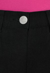 Zwarte katoenen broek met een zilverkleurige metalen knoop met gegraveerde details aan de taille, gecombineerd met een roze top. Close-up weergave.