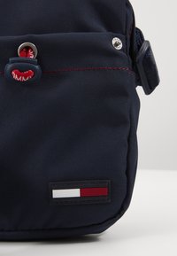 Borsa a spalla in tessuto blu navy con cuciture rosse, dotata di zip, di una linguetta in gomma e di un'etichetta rettangolare con logo bianco e accenti rossi.