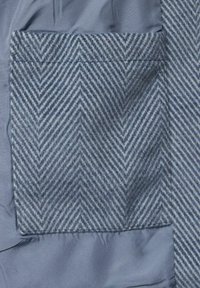 Blått tyg med en ficka i fiskbensmönster. Fickan är gjord av en texturerad ullblandning, vilket kontrasterar mot det släta tyget.