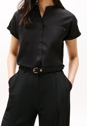 Mujer con una camisa negra de manga corta abotonada con bolsillos delanteros, metida dentro de pantalones negros de cintura alta con un cinturón negro.