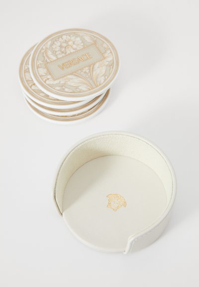 Podtácky Versace s květinovým motivem v krémové a zlaté barvě, navrstvené vedle kulatého bílého koženého držáku se zlatým logem.