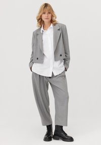 Szary krótki żakiet na białej koszuli zapinanej na guziki, zestawiony z luźnymi szarymi spodniami i czarnymi sznurowanymi butami. Gładka tkanina o strukturalnym kroju.