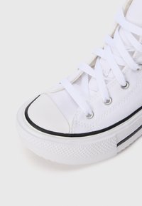 Sapatilha alta de lona branca com atacadores brancos, ilhós metálicos, detalhes em preto e sola de borracha texturizada, sobre um fundo claro.