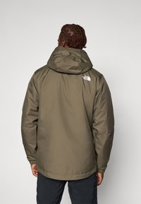 Veste imperméable vert olive avec capuche, manches élastiques et bas ajustable ; présente un logo blanc sur l'épaule gauche.