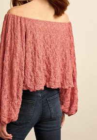Off-the-shoulder, struktureret top i støvet rose med luftige ærmer; parret med mørke, tætsiddende jeans med baglommer.
