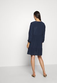 Mörkblå klänning med långa ärmar, figurnära liv och lager på lager volangkjol. Slät tyg med en något løs passform baktill.