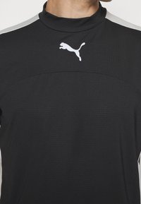 Camisa atlética negra con cuello alto, que presenta un logo blanco de Puma y un sutil patrón texturizado en la tela. Detalles blancos en los hombros.