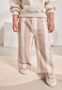 Pantalones de rayas beige y crema fabricados con tela texturizada, combinados con zapatos de beige claro que cuentan con dos correas de velcro.