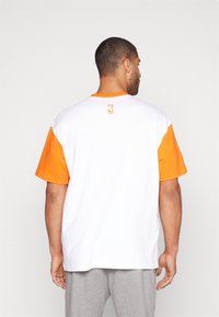 Rövid ujjú póló, fehér alapszíne narancssárga ujjakkal és gallérral. Hátul, a nyaknál egy kis NBA-logo látható.