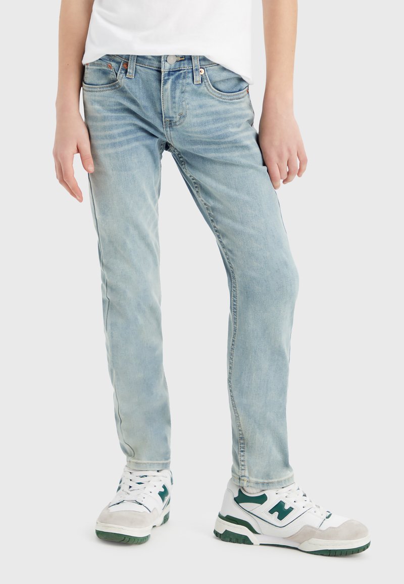 Jeans de mezclilla azul claro con un corte ajustado, desvanecimiento sutil, diseño clásico de cinco bolsillos y costuras blancas, combinados con zapatillas atléticas blancas y verdes.