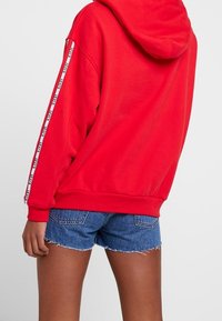 Red hoodie with a drawstring and white side stripes featuring branded text. Paired with blue denim shorts with frayed edges.