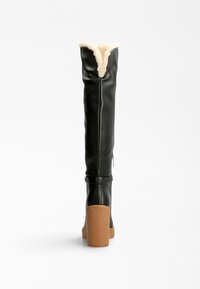 Bottes en cuir noir montant au-dessus du genou avec une doublure en fausse fourrure crème, dotées d'un talon bloc marron texturé et d'une fermeture éclair latérale.