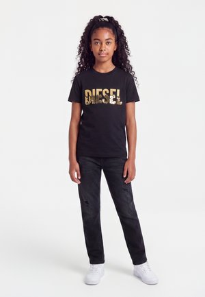 Fille aux longs cheveux bouclés portant un t-shirt Diesel noir à sequins, un jean noir déchiré et des baskets blanches, debout devant un fond blanc.