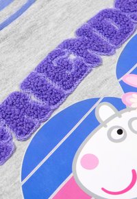 Tessuto grigio con lettere testurizzate viola, caratterizzato dal volto di un personaggio dei cartoni animati in un design circolare blu e rosa. Accenti brillanti e colorati.